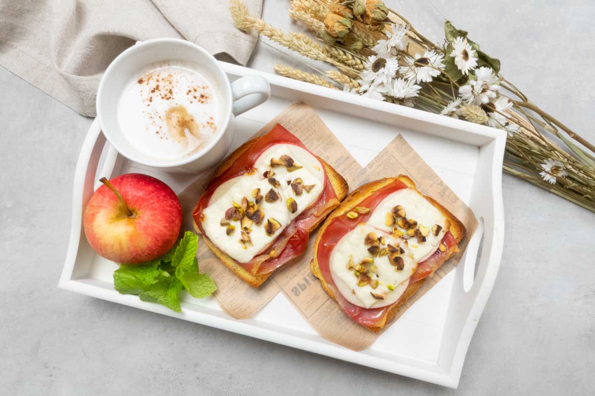 Foto cenital de unas tostadas gratinadas de jamón serrano con manzana servidas con café con leche en una bandeja blanca de madera / Foto de María Alcázar