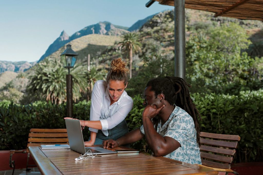 Dos personas colaborando frente a un portátil en una mesa de madera, en una terraza rodeada de vegetación y montañas al fondo, bajo una pérgola